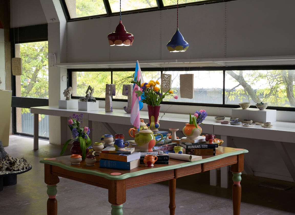 Wooden table with stacks of books, ceramic vessels, flowers, jewelry, and a flag.