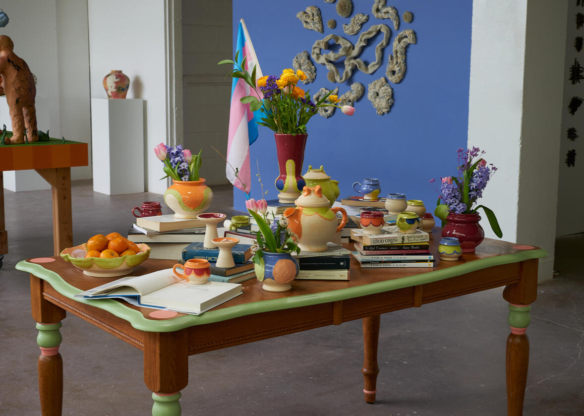 Wooden table with stacks of books, ceramic vessels, flowers, jewelry, and a flag.