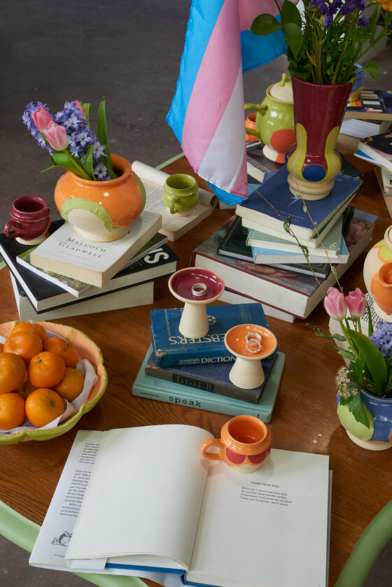 Detail photo of mixed media art installation showing stacks of books, ceramic vessels, oranges, flowers, and silver rings.